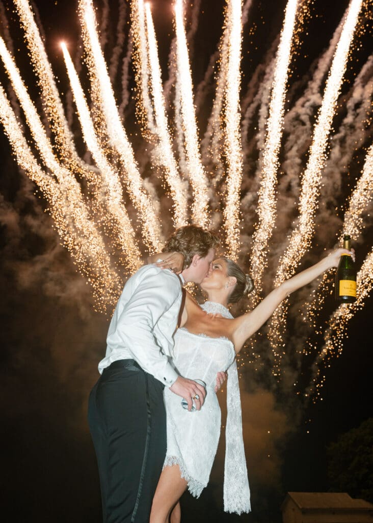 Bride and groom kissing with veuvre criquot while fireworks explode in the background creating a grand celebration